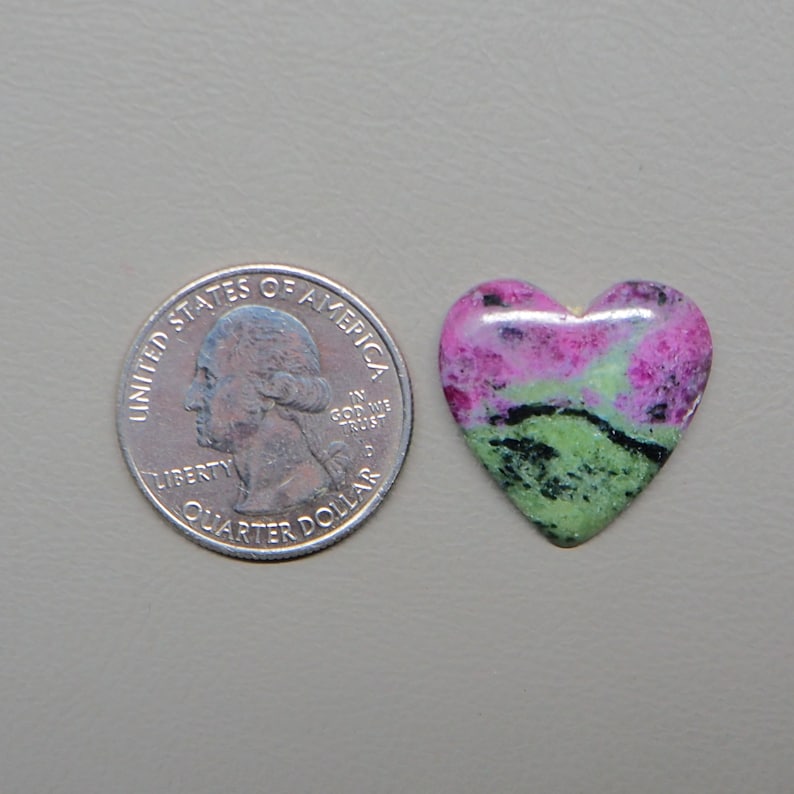 May include: A heart-shaped gemstone with a quarter coin next to it for size comparison. The gemstone features a mix of pink, green, and black colors. The coin is a United States quarter dollar.
