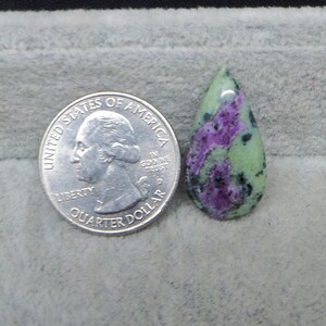 May include: A teardrop-shaped gemstone with a light green base, vibrant purple and black inclusions, next to a U.S. quarter for size comparison. The polished stone has a smooth, glossy surface, showcasing natural patterns and colors.