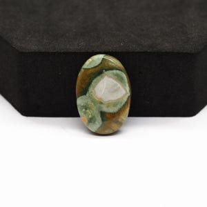 K&ouml;nnte beinhalten: Ein ovaler Cabochon-Edelstein mit einer polierten Oberfl&auml;che und einem gesprenkelten Muster aus erdigem Braun, Salbeigr&uuml;n und cremefarbenen Einschl&uuml;ssen. Der Stein zeigt eine glatte, abgerundete Oberseite und einen markanten, wei&szlig;en Fleck.