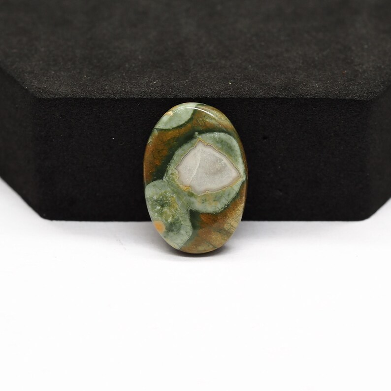 K&ouml;nnte beinhalten: Ein ovaler Cabochon-Edelstein mit einer polierten Oberfl&auml;che und einem gesprenkelten Muster aus erdigem Braun, Salbeigr&uuml;n und cremefarbenen Einschl&uuml;ssen. Der Stein zeigt eine glatte, abgerundete Oberseite und einen markanten, wei&szlig;en Fleck.