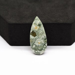K&ouml;nnte beinhalten: Ein tropfenf&ouml;rmiger Cabochon-Edelstein mit einem gesprenkelten Muster aus Salbeigr&uuml;n, Oliv und Wei&szlig;. Der Stein hat eine glatte, polierte Oberfl&auml;che mit einem deutlichen dunkelbraunen Einschluss nahe der Spitze und einem spitz zulaufenden Ende.