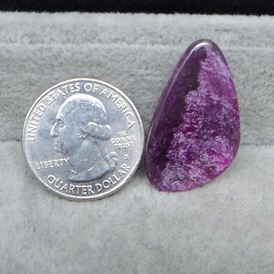 May include: A polished, teardrop-shaped gemstone with a vibrant purple hue and a textured surface. A United States quarter dollar coin is next to the gemstone for size comparison.