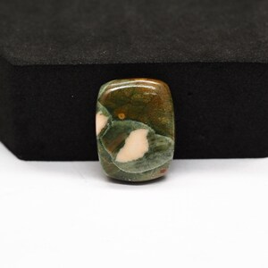 K&ouml;nnte beinhalten: Ein rechteckiger, polierter Edelstein-Cabochon mit einem gesprenkelten Muster aus tiefem Waldgr&uuml;n, erdigem Braun und cremewei&szlig;en Flecken. Der Stein hat eine glatte, gl&auml;nzende Oberfl&auml;che mit sichtbarer nat&uuml;rlicher Maserung.