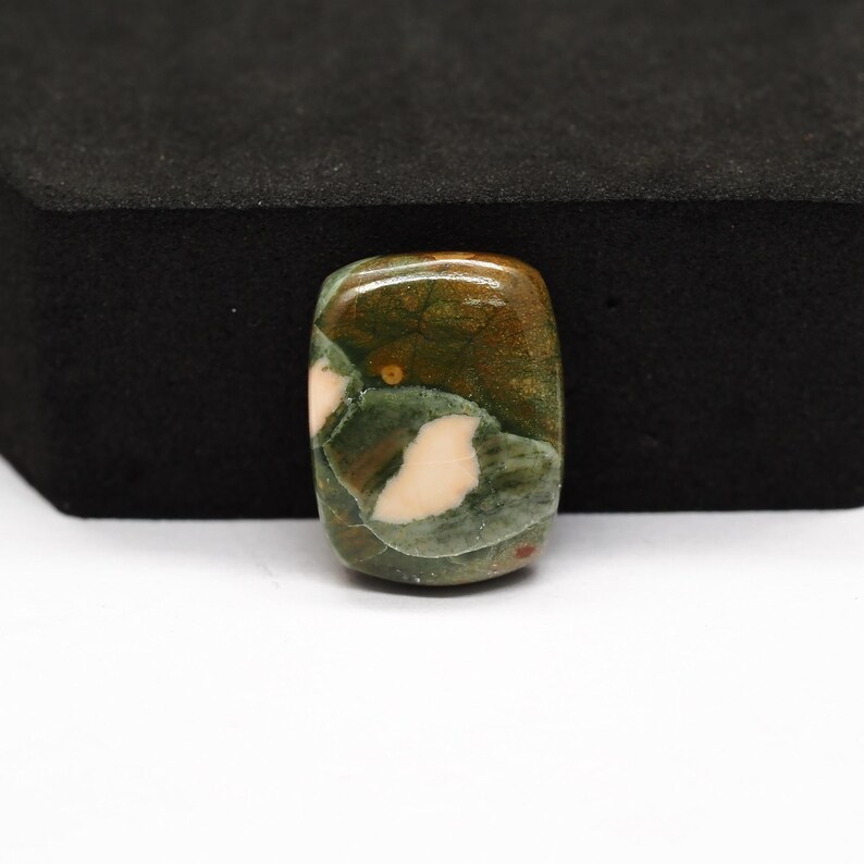 K&ouml;nnte beinhalten: Ein rechteckiger, polierter Edelstein-Cabochon mit einem gesprenkelten Muster aus tiefem Waldgr&uuml;n, erdigem Braun und cremewei&szlig;en Flecken. Der Stein hat eine glatte, gl&auml;nzende Oberfl&auml;che mit sichtbarer nat&uuml;rlicher Maserung.