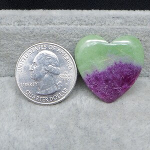 May include: A heart-shaped stone with a green and purple color gradient. A U.S. quarter coin is next to the stone for size comparison. The stone is smooth and polished, with a glossy finish. The heart is resting on a gray surface.