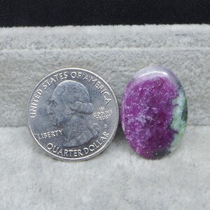 May include: An oval-shaped gemstone with a vibrant mix of colors, including deep magenta and green, is displayed next to a U.S. quarter for size comparison. The gemstone's polished surface reflects light, showcasing its natural patterns and textures.