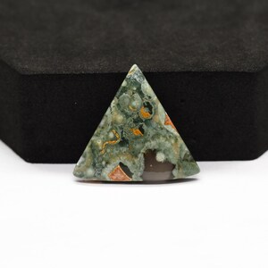 K&ouml;nnte beinhalten: Ein dreieckiger, polierter Edelstein-Cabochon mit einem gesprenkelten Muster aus Salbeigr&uuml;n, dunklem Waldgr&uuml;n und cremefarbenen kreisf&ouml;rmigen Einschl&uuml;ssen, akzentuiert durch kleine Flecken in gebranntem Orange und eine glatte, dunkelbraune Fl&auml;che.