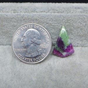 May include: A triangular-shaped gemstone with a mix of green, white, and magenta colors. A U.S. quarter coin is next to the gemstone for size comparison. The gemstone has a polished surface and a natural, organic appearance.