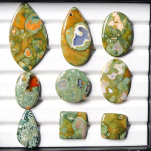 以下が含まれることがあります： 楕円形、涙型、正方形など、さまざまな形状の9つの研磨された宝石のコレクション。石は、独特のパターンとテクスチャを備えた、緑、オレンジ、茶色の色の混合物を特徴としています。宝石は黒いトレイに表示されます。