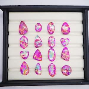 May include: A collection of polished gemstones in various shapes, including ovals, teardrops, and hearts. The stones feature a vibrant mix of pink, purple, blue, and gold colors, set against a white background within a black display tray.
