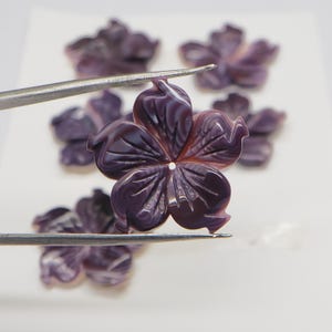 May include: Close-up of several carved purple flower-shaped beads. One bead is held by tweezers, showcasing the intricate petal details and a central hole. The beads have a glossy finish and are arranged on a white surface.