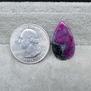 May include: A teardrop-shaped gemstone with a vibrant mix of colors, including deep magenta, black, and hints of green. A U.S. quarter coin is placed next to the gemstone for size comparison. The gemstone has a polished surface.