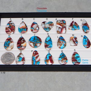 May include: A collection of polished gemstone cabochons in various shapes, including teardrops, ovals, and a heart. The stones feature a mix of turquoise, chocolate brown, and white colors, with metallic inclusions. The gemstones are displayed in a black tray.