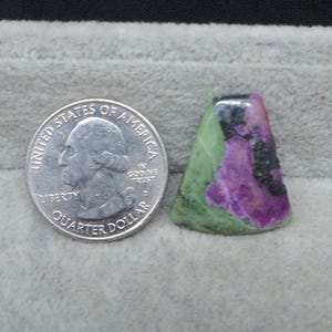 May include: A polished gemstone with a mix of colors, including green, purple, and black. The stone is next to a quarter dollar coin for size comparison. The gemstone has a unique shape and texture.
