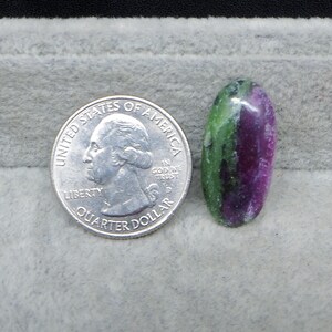 May include: An oval-shaped ruby zoisite gemstone with green and magenta hues. A U.S. quarter dollar coin is next to the gemstone for size comparison. The gemstone is set against a gray background.