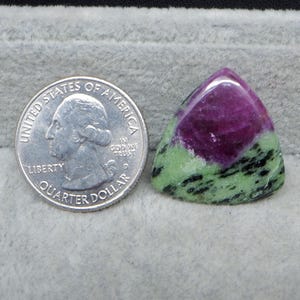 May include: A polished, triangular gemstone with a vibrant color combination. The top portion is a deep magenta, transitioning to a light green base with black inclusions. A US quarter coin is next to the gemstone for size comparison.