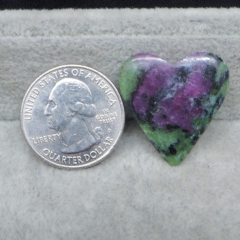 May include: A heart-shaped gemstone with a mix of green, purple, and black colors. A US quarter coin is next to the gemstone for size comparison. The gemstone is polished and smooth.