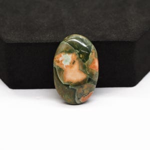 K&ouml;nnte beinhalten: Ein ovaler Cabochon-Edelstein mit einer polierten Oberfl&auml;che, die ein gesprenkeltes Muster aus tiefem Waldgr&uuml;n, Oliv und weichen pfirsichfarbenen Einschl&uuml;ssen aufweist. Der Stein hat eine glatte, abgerundete W&ouml;lbung und eine gl&auml;nzende Oberfl&auml;che.