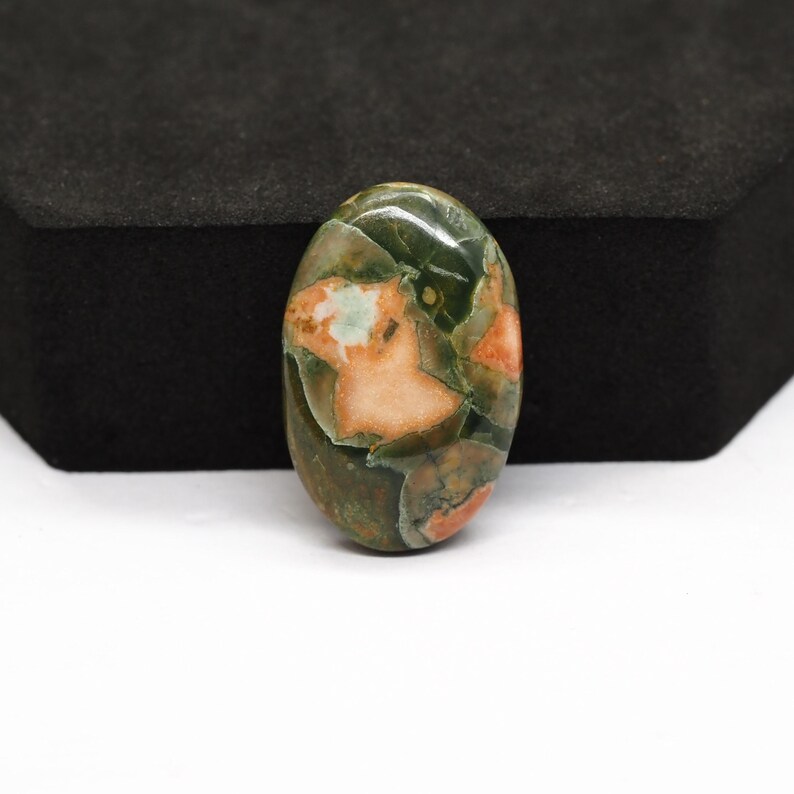 K&ouml;nnte beinhalten: Ein ovaler Cabochon-Edelstein mit einer polierten Oberfl&auml;che, die ein gesprenkeltes Muster aus tiefem Waldgr&uuml;n, Oliv und weichen pfirsichfarbenen Einschl&uuml;ssen aufweist. Der Stein hat eine glatte, abgerundete W&ouml;lbung und eine gl&auml;nzende Oberfl&auml;che.