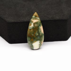 K&ouml;nnte beinhalten: Ein tropfenf&ouml;rmiger, polierter Edelstein-Cabochon mit einem gesprenkelten Muster in Waldgr&uuml;n, Olivbraun und Cremewei&szlig;. Der Stein hat eine glatte, gl&auml;nzende Oberfl&auml;che mit einer abgerundeten Spitze und einem spitz zulaufenden Ende mit nat&uuml;rlichen Einschl&uuml;ssen.