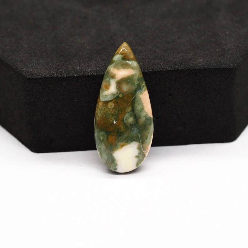K&ouml;nnte beinhalten: Ein tropfenf&ouml;rmiger, polierter Edelstein-Cabochon mit einem gesprenkelten Muster in Waldgr&uuml;n, Olivbraun und Cremewei&szlig;. Der Stein hat eine glatte, gl&auml;nzende Oberfl&auml;che mit einer abgerundeten Spitze und einem spitz zulaufenden Ende mit nat&uuml;rlichen Einschl&uuml;ssen.