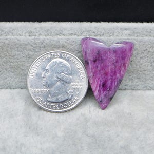 May include: A heart-shaped, polished, purple gemstone with black inclusions. The gemstone is next to a United States quarter for size comparison. The stone has a smooth, glossy finish and a unique, natural pattern.