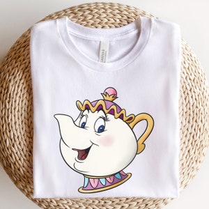 Op de afbeelding: Wit T-shirt met een cartoon afbeelding van Mrs. Potts, het theepotkarakter uit Belle en het Beest. Ze heeft een roze en gele hoed en een blauwe en witte rand rond haar basis.