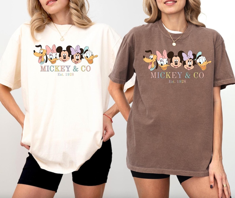 May include: Two women wearing t-shirts with a Mickey Mouse and friends graphic. The shirts are in cream and brown colors. The text on the shirts reads "Mickey & Co. Est. 1928".