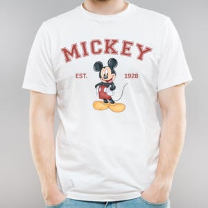 May include: White t-shirt with a red and black Mickey Mouse graphic. The text "Mickey" is in a red arch above the graphic. The text "Est. 1928" is in red below the graphic.