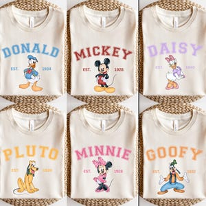 May include: Six cream-colored t-shirts with different Disney character designs. Each shirt features a character's name in a colorful, varsity-style font, along with the character's established year. The characters featured are Donald Duck, Mickey Mouse, Daisy Duck, Pluto, Minnie Mouse, and Goofy.