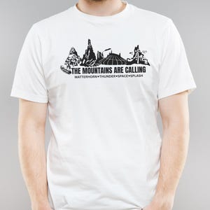 以下が含まれることがあります： 黒い山のシルエットのグラフィックと「THE MOUNTAINS ARE CALLING」のテキストがプリントされた白いTシャツ。「MATTERHORN THUNDER SPACE SPLASH」も含まれています。 このシャツは半袖で、クルーネックです。
