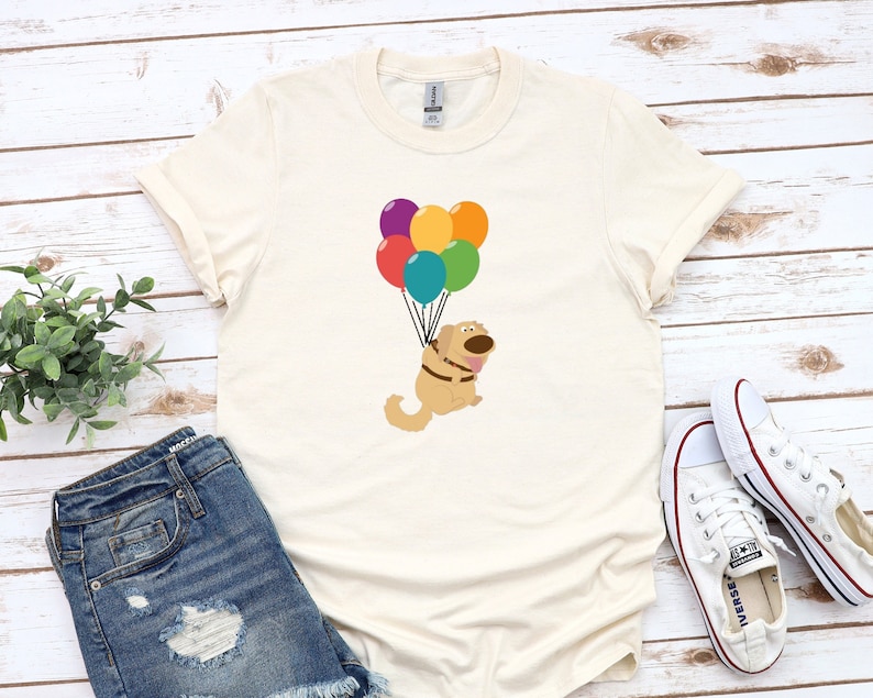 May include: A beige t-shirt with a cartoon dog holding onto colorful balloons. The dog is brown with a black nose and eyes. The balloons are red, orange, yellow, green, blue, and purple.