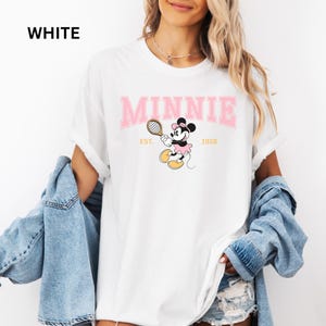 May include: White t-shirt with the word "MINNIE" in pink block letters above a cartoon Minnie Mouse playing tennis. The text "EST. 1928" is below the graphic. The word "WHITE" is printed in black in the upper left corner.