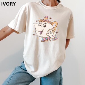 Puede incluir: Camiseta de color marfil con un diseño de dibujos animados de tetera y taza. El gráfico muestra una tetera sonriente con un asa dorada y una taza, rodeada de estrellas. La palabra "IVORY" está impresa en la parte superior.