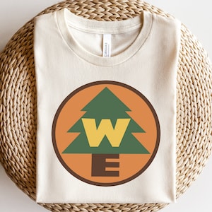 May include: A white t-shirt with a brown, orange, and green circular design featuring a stylized pine tree and the letters "WE".