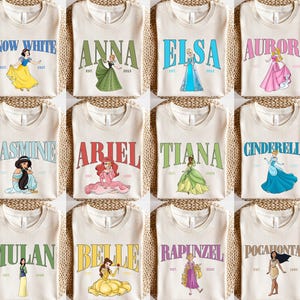 May include: Twelve white t-shirts with different Disney princess characters printed on them. Each shirt features a princess's name, a stylized 'EST' for established, and a year. The princesses featured are Snow White, Anna, Elsa, Aurora, Jasmine, Ariel, Tiana, Cinderella, Mulan, Belle, Rapunzel, and Pocahontas.