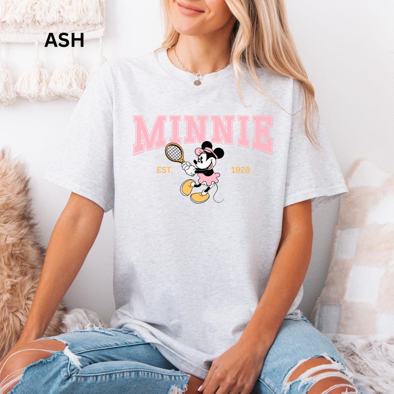 May include: Ash gray t-shirt featuring a pink "MINNIE" arched text design with a Minnie Mouse graphic playing tennis. The graphic includes the text "EST. 1928". The shirt is a light gray color.