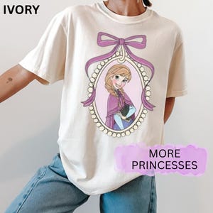 May include: Cream-coloured t-shirt with a cartoon princess image in an oval frame, decorated with a purple bow and ribbon. The text "MORE PRINCESSES" is on a purple banner. The shirt is made of a soft material.