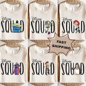 Puede incluir: Camisetas color crema con la frase "Disney Squad" en negro, cada una con un personaje diferente de Toy Story: un extraterrestre, Buzz Lightyear, Jessie, Zurg, Woody y un pingüino. Las camisetas se exhiben sobre una superficie tejida.