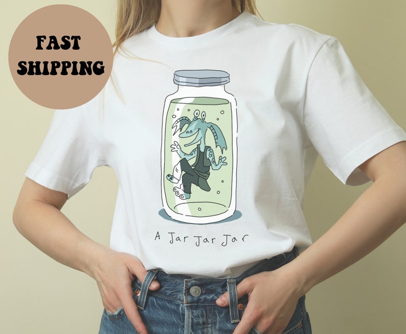 Puede incluir: Camiseta blanca con una ilustraci&oacute;n de dibujos animados de una criatura azul dentro de un tarro. El texto "A Jar Jar Jar" est&aacute; impreso debajo. El dise&ntilde;o es de estilo caricatura.