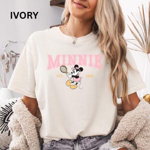 May include: Ivory-colored t-shirt featuring the word "MINNIE" in pink block letters above a cartoon Minnie Mouse playing tennis. The shirt also includes the text "EST. 1928".