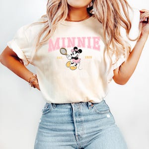 May include: Cream-colored t-shirt featuring the word "MINNIE" in pink block letters above a cartoon Minnie Mouse playing tennis. The graphic includes the text "EST. 1928". The shirt is paired with blue jeans.