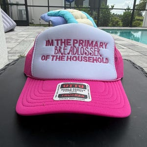 May include: A bright pink and white trucker hat featuring a foam front panel with the embroidered text "IM THE PRIMARY BREADLOSSER OF THE HOUSEHOLD" in pink thread. The hat includes a pink mesh back, a curved brim, and an Otto brand sticker on the front.