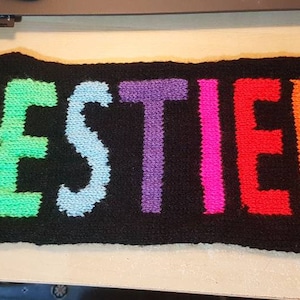 May include: A black knitted scarf with the word "DESTIEL" in rainbow-colored letters. The letters are yellow, green, blue, purple, pink, red, and orange. The scarf is rectangular.