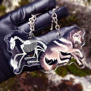 May include: A clear acrylic keychain with a wolf skeleton on the left side and a wolf with fur on the right side. The keychain is in the shape of a wolf. The wolf skeleton is white and the wolf with fur is gray and white. The keychain has a silver metal chain attached to it.