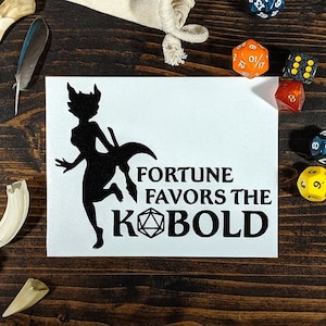 May include: A white card with a black silhouette of a kobold holding a dagger. The text "FORTUNE FAVORS THE KOBOLD" is printed below the silhouette. The card is surrounded by dice and other gaming accessories on a wooden table.