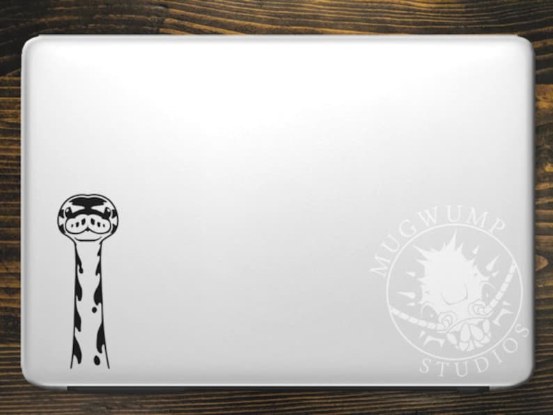 May include: Black and white sticker of a snake's head and neck on a silver laptop. The sticker is on the left side of the laptop. On the right side of the laptop is a white sticker of a logo that says "Mugwump Studios".