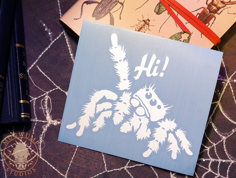 A photograph of a cute jumping spider decal. The jumping spider is looking up and raising one leg to wave a greeting. He says 'Hi!' The photograph is of the decal sitting top of a spiderweb lace tablecloth and a bug-themed notebook.