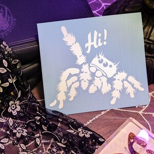 A photograph of a cute jumping spider decal. The jumping spider is looking up and raising one leg to wave a greeting. He says 'Hi!' The photograph is of the decal sitting top of a spiderweb lace tablecloth.