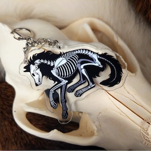 Peut inclure: Un porte-cl&eacute;s en acrylique noir et blanc repr&eacute;sentant un loup squelette avec une silhouette noire. Le porte-cl&eacute;s est attach&eacute; &agrave; une cha&icirc;ne argent&eacute;e.